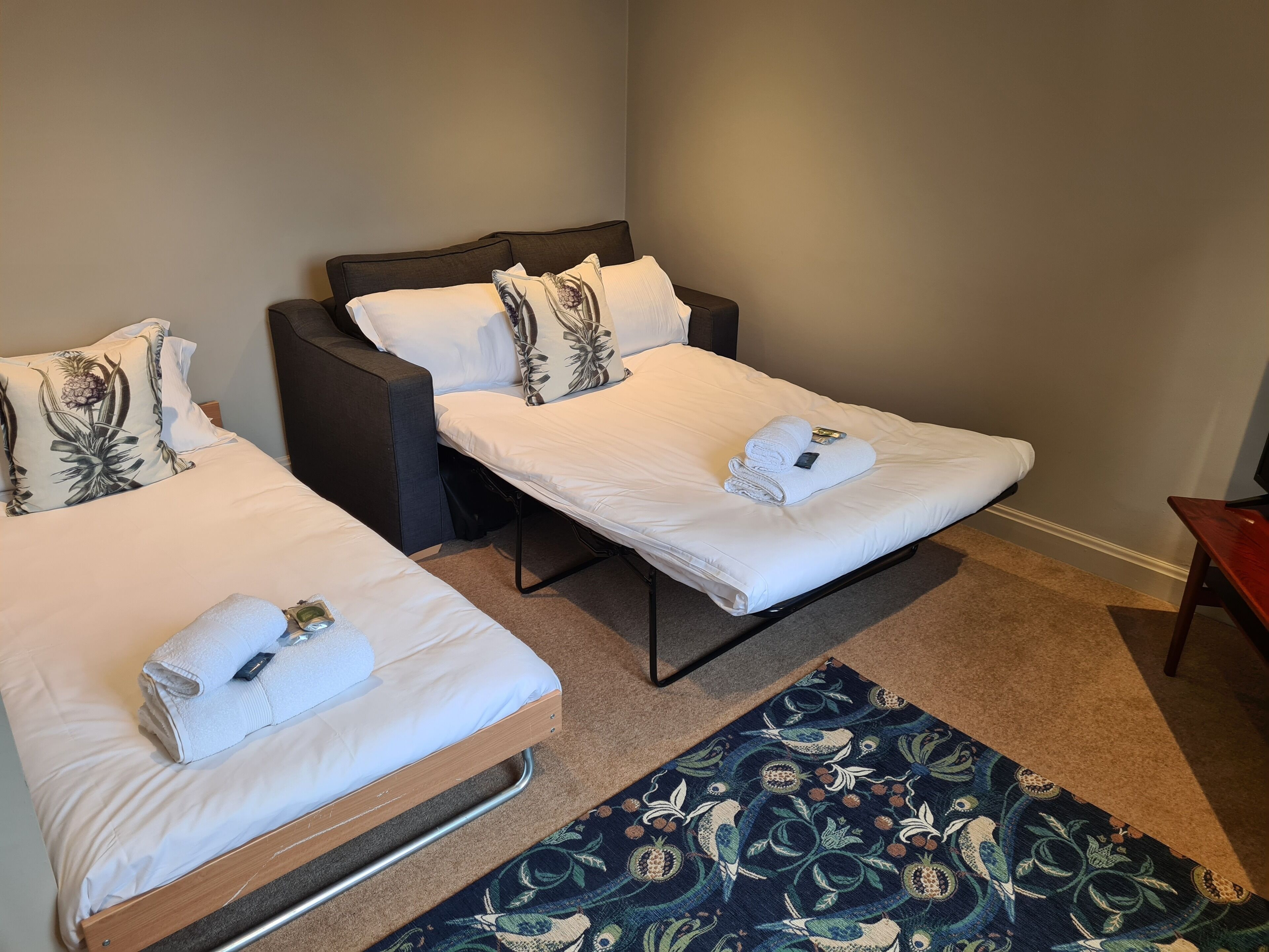 A hotel room with two single beds, each with a white mattress and matching pillows, a dark headboard, and a blue patterned rug on the floor.