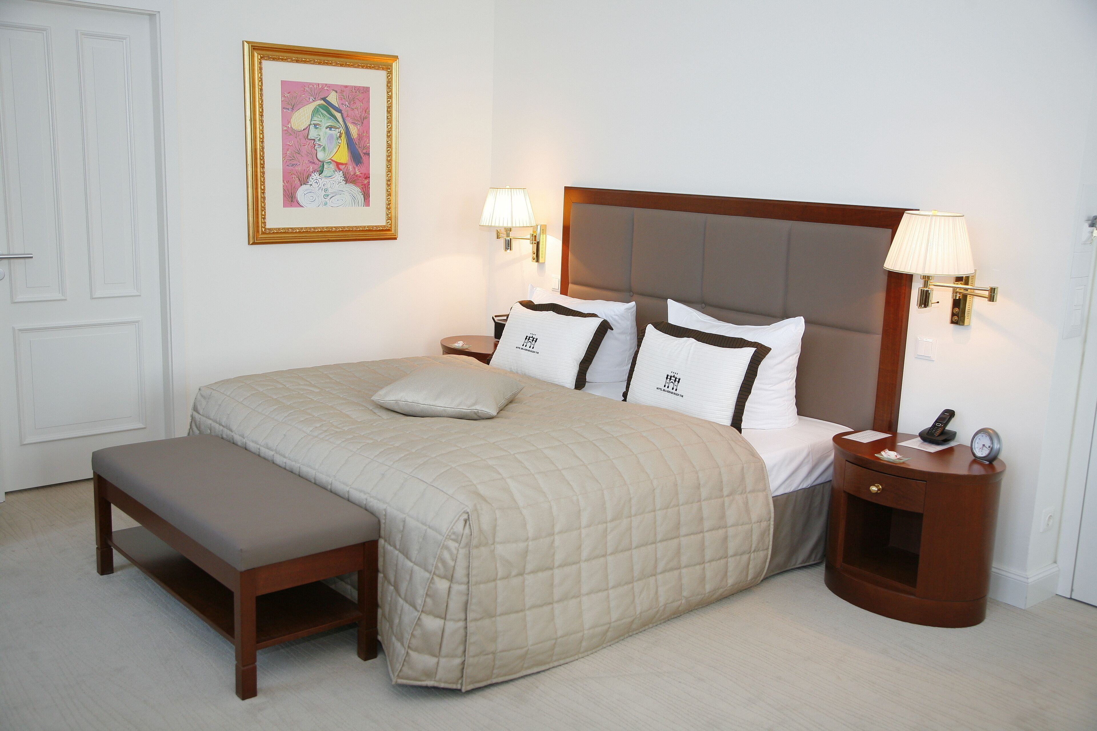 Un dormitorio ordenado con cama, mesitas de noche, un banco y una obra de arte enmarcada en la pared.