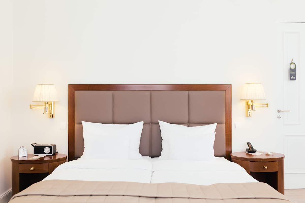 Cama bem arrumada com cabeceira de madeira, duas mesas de cabeceira com abajures e um relógio em uma das mesas.