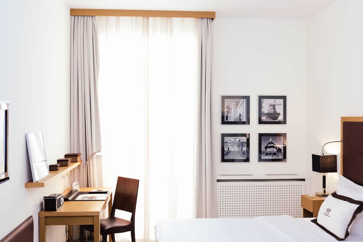 Quarto de hotel com cama, escrivaninha com cadeira, televisão e radiador. Há quatro quadros na parede.