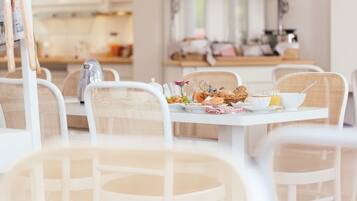 Un comedor con una mesa blanca dispuesta para el desayuno, sillas de mimbre y una cocina al fondo.
