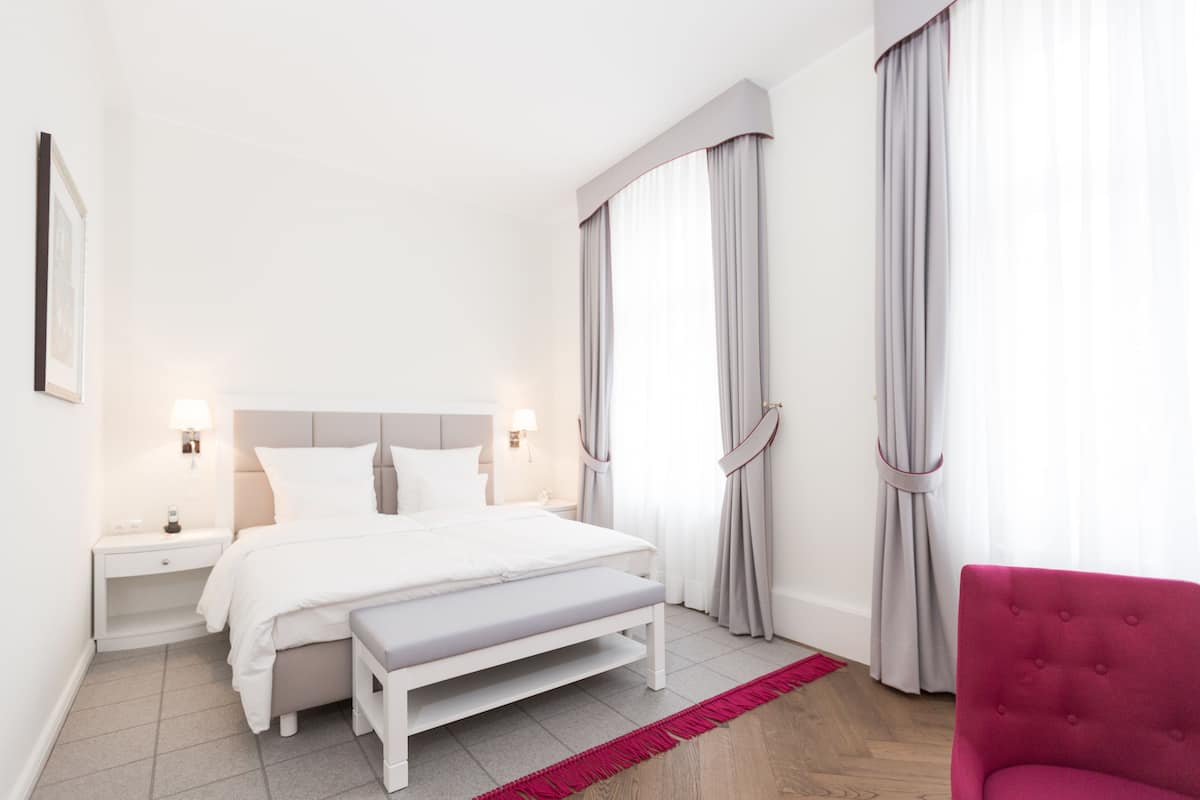 Quarto de hotel moderno com uma cama grande, mesas de cabeceira, um banco e uma cadeira vermelha.