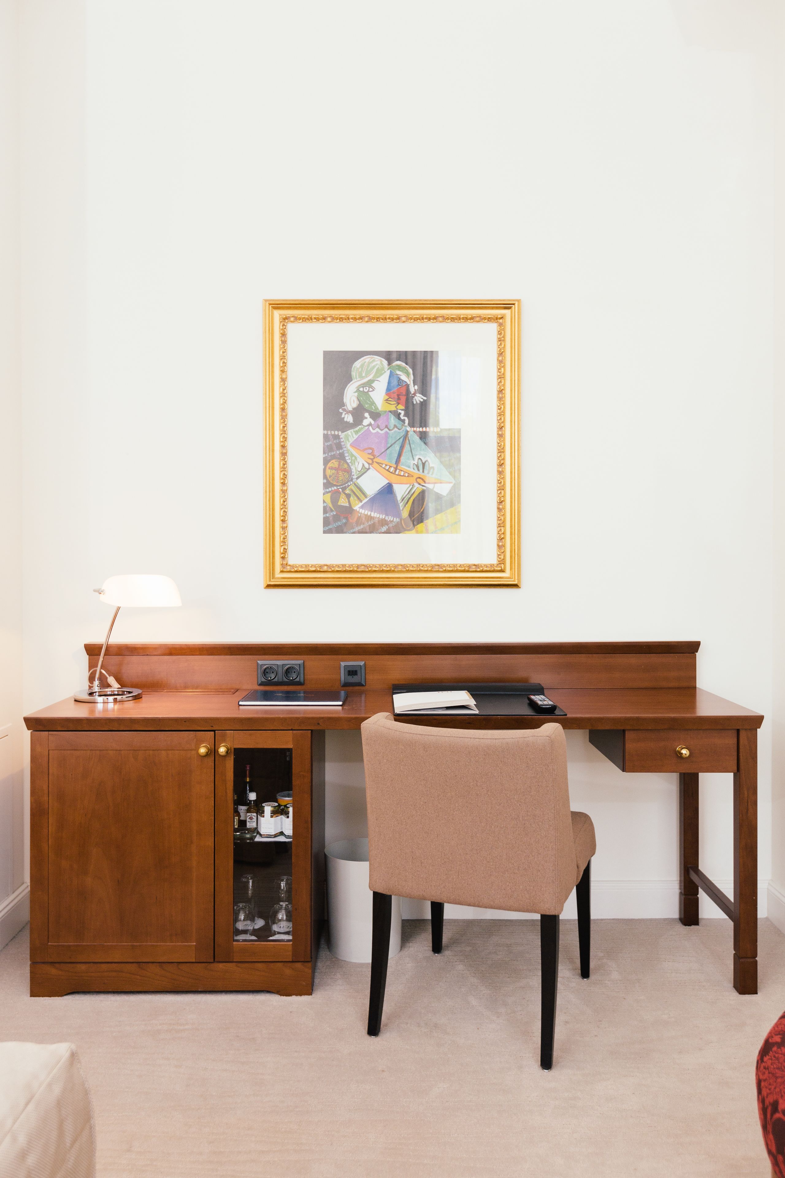 A wooden desk with a chair, a framed artwork on the wall, and a cabinet with glass doors.