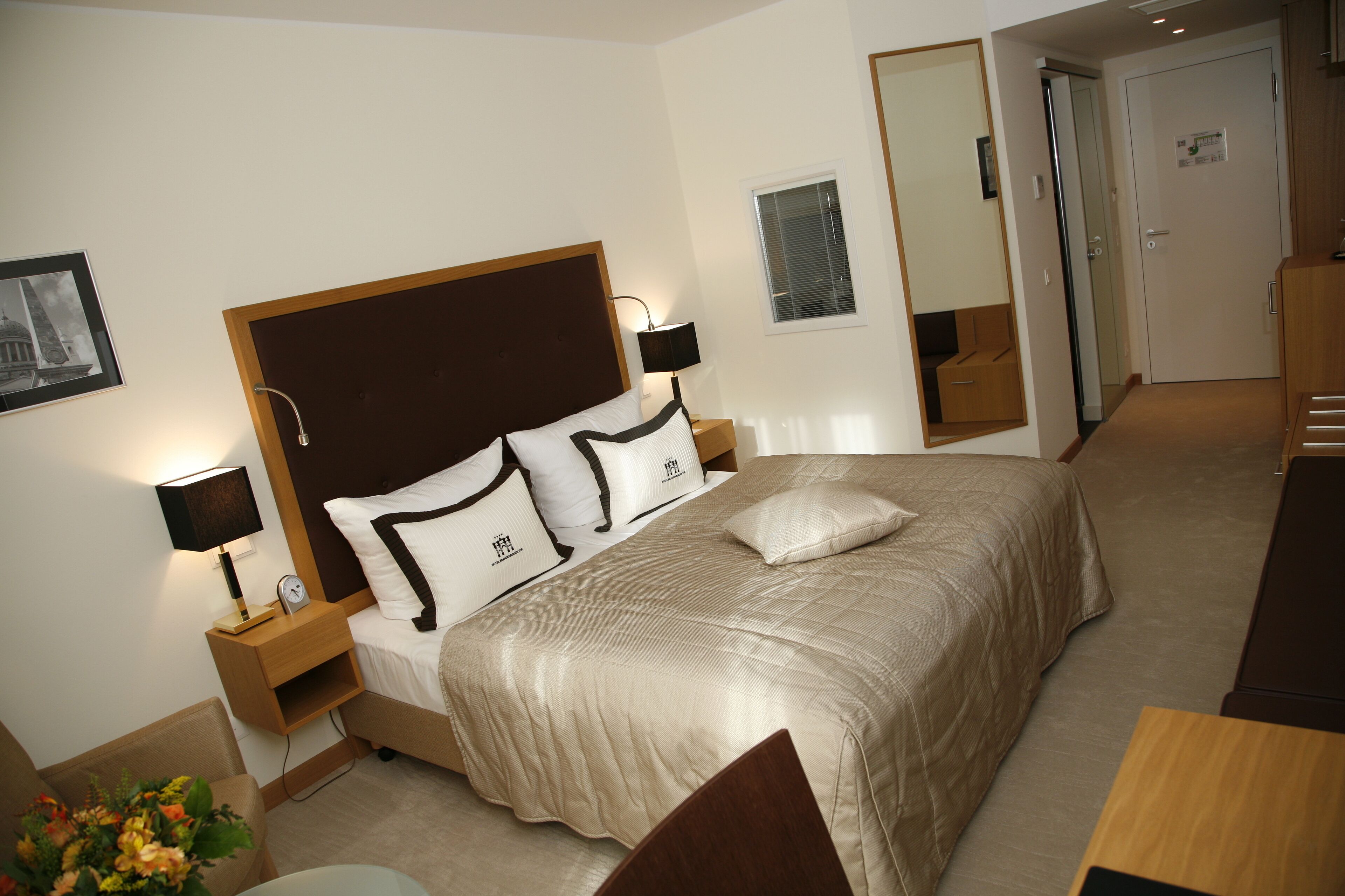 A hotel room with a large bed, a nightstand with a lamp, a bedside table, a mirror, and a door.