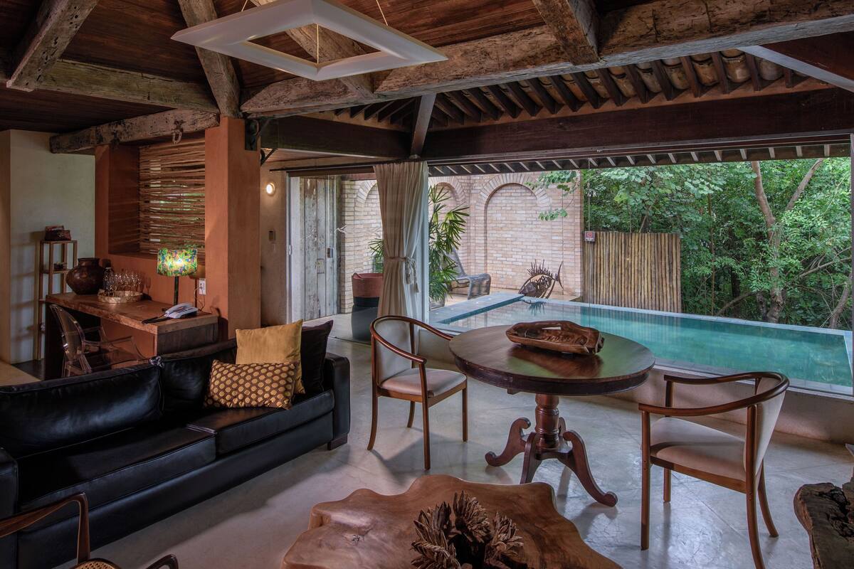 A cozy living area with a black leather sofa, wooden dining table, and chairs, a small kitchen area, and a glass-enclosed outdoor swimming pool.