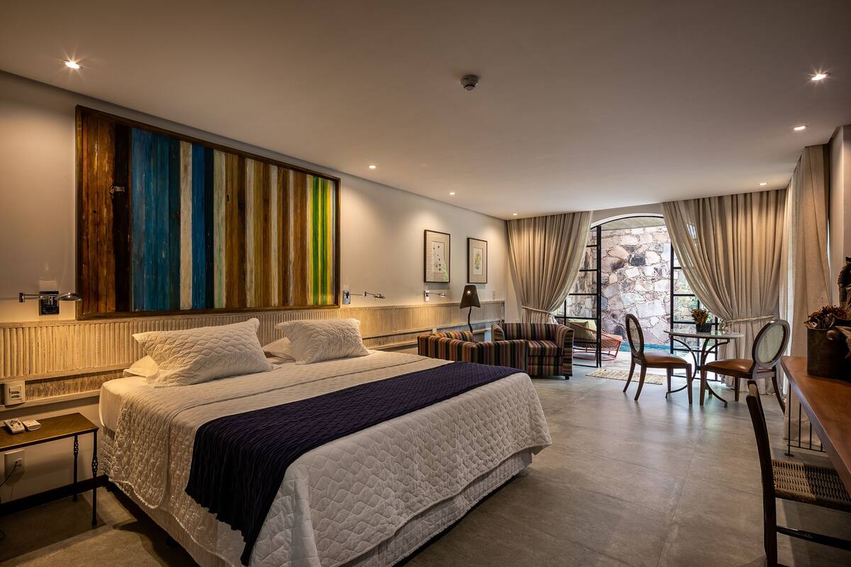 A modern hotel room with a large bed, a seating area with a table and chairs, and a view of the outdoors through a window.