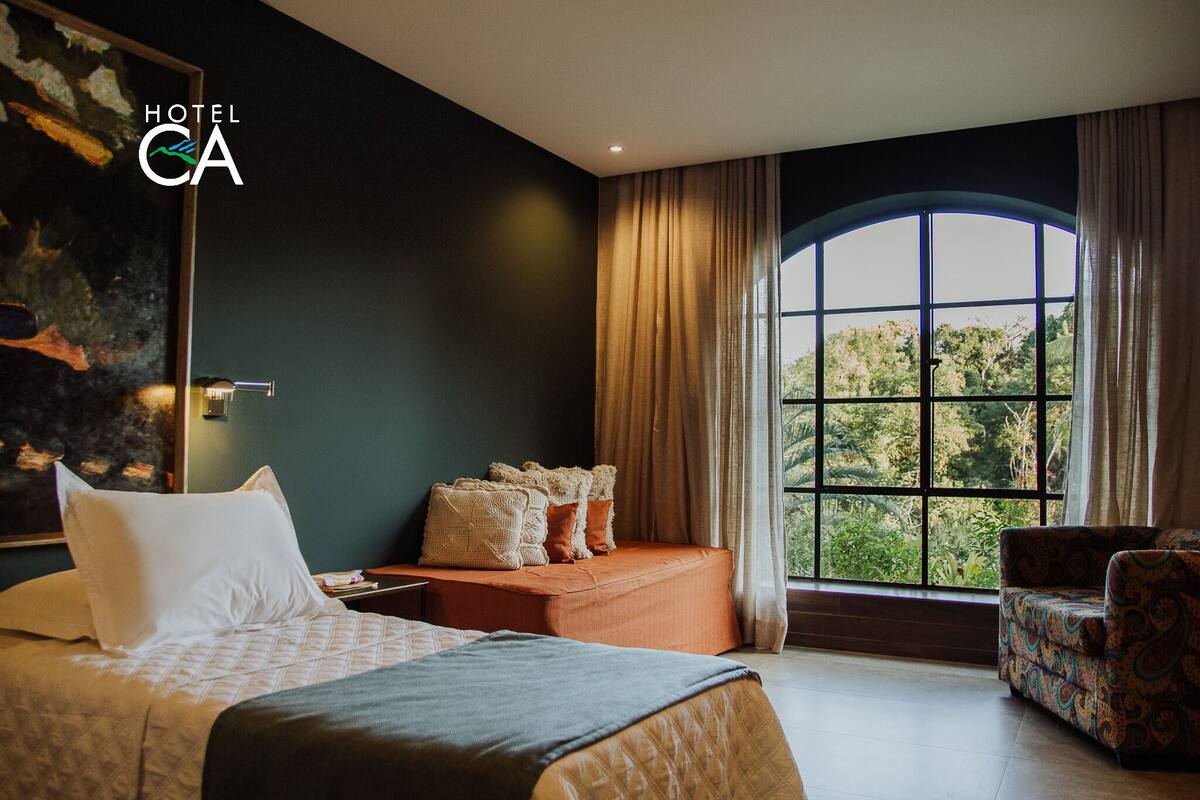 A hotel room with a large bed, a sofa, a chair, and a window with a view of trees.