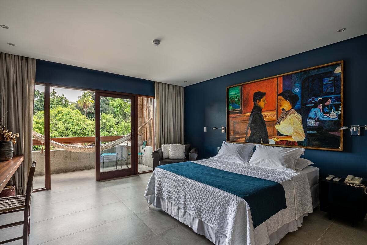 A bedroom with a large bed, a painting, a desk, and a view of greenery.