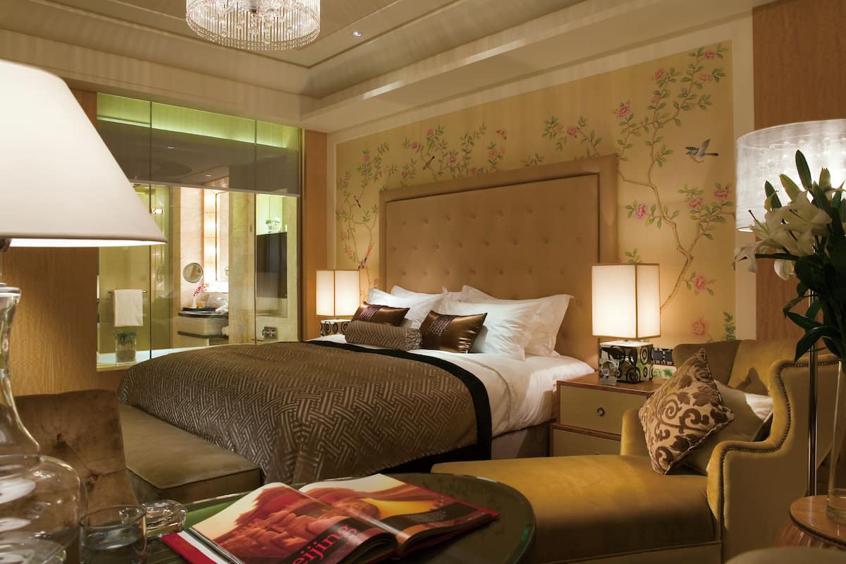 A hotel room with a large bed, two bedside tables, a sitting area with a chair, a table with a magazine, and a vase with flowers.