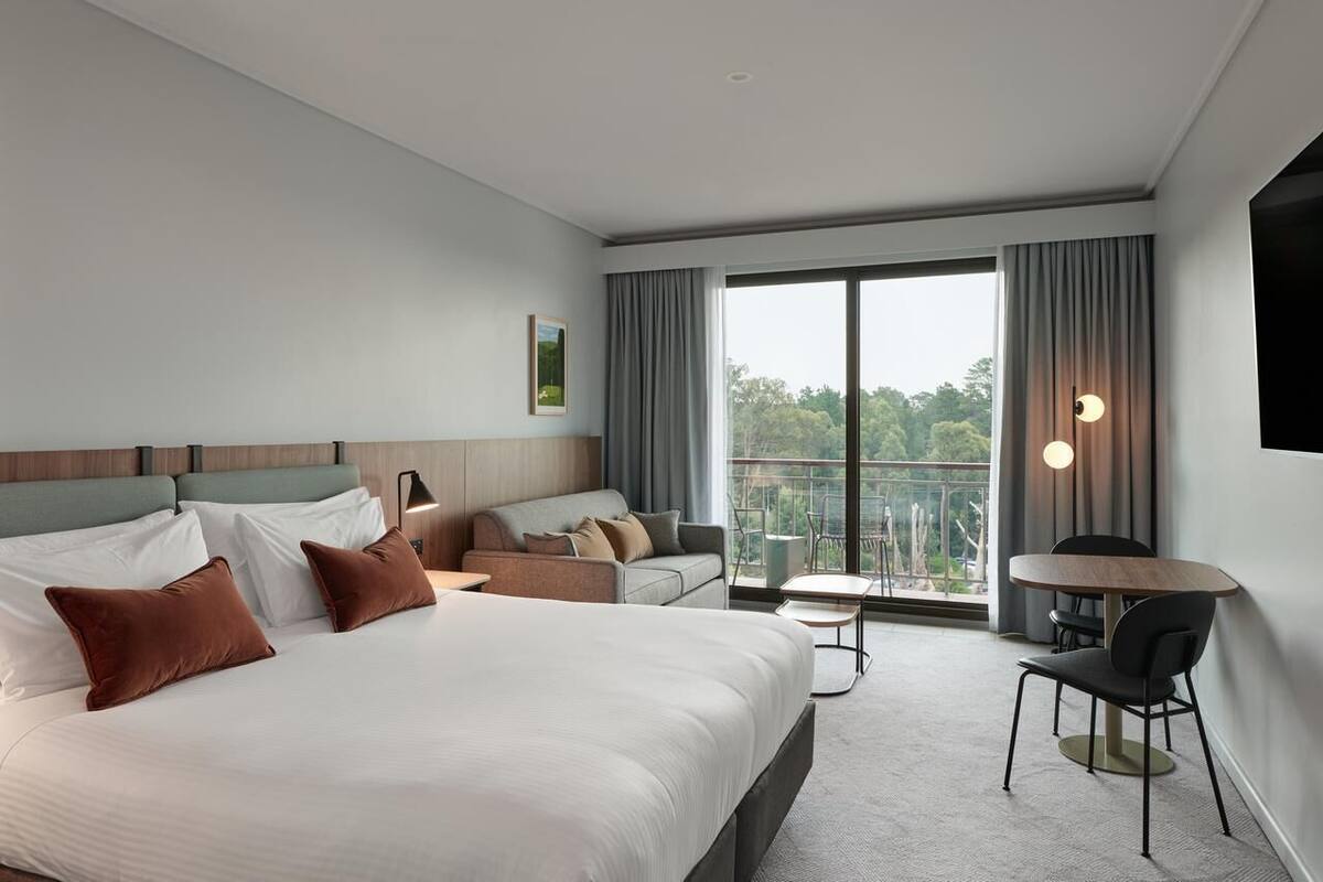 A modern hotel room with a large bed, a sofa, a small table, and a balcony with a view.