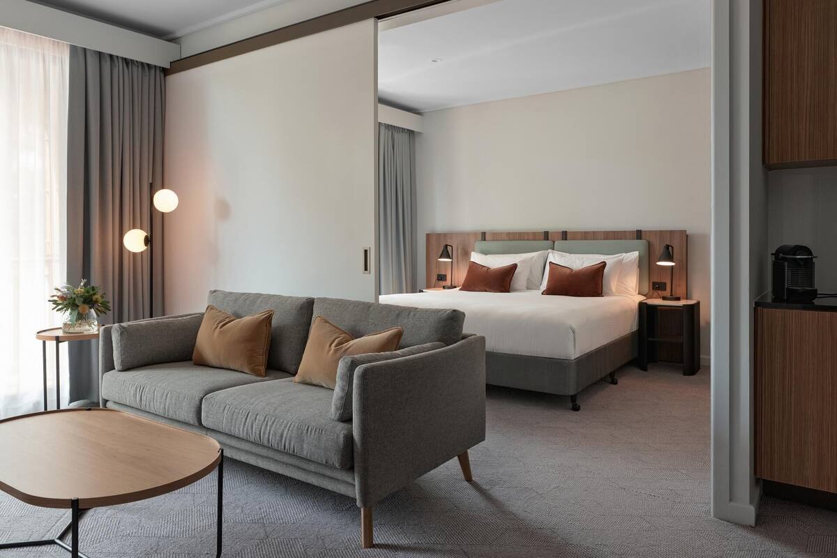 A hotel room with a sofa, a small table, a bed with pillows, a nightstand with a lamp, and a wooden cabinet.