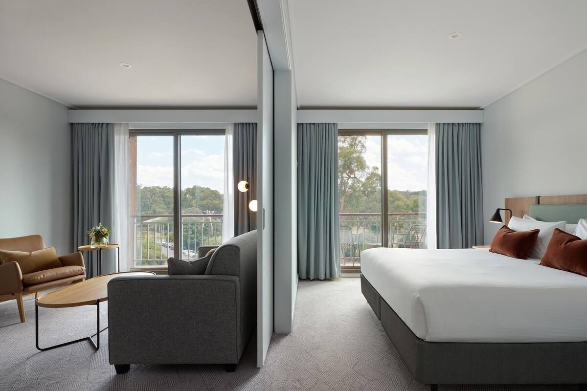 A modern hotel room with a large bed, a sofa, a small table, and a balcony with a view of the city.