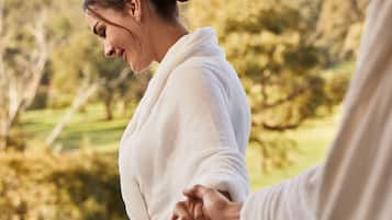 A couple in white bathrobes holding hands on a balcony with a view of a green landscape and hills.