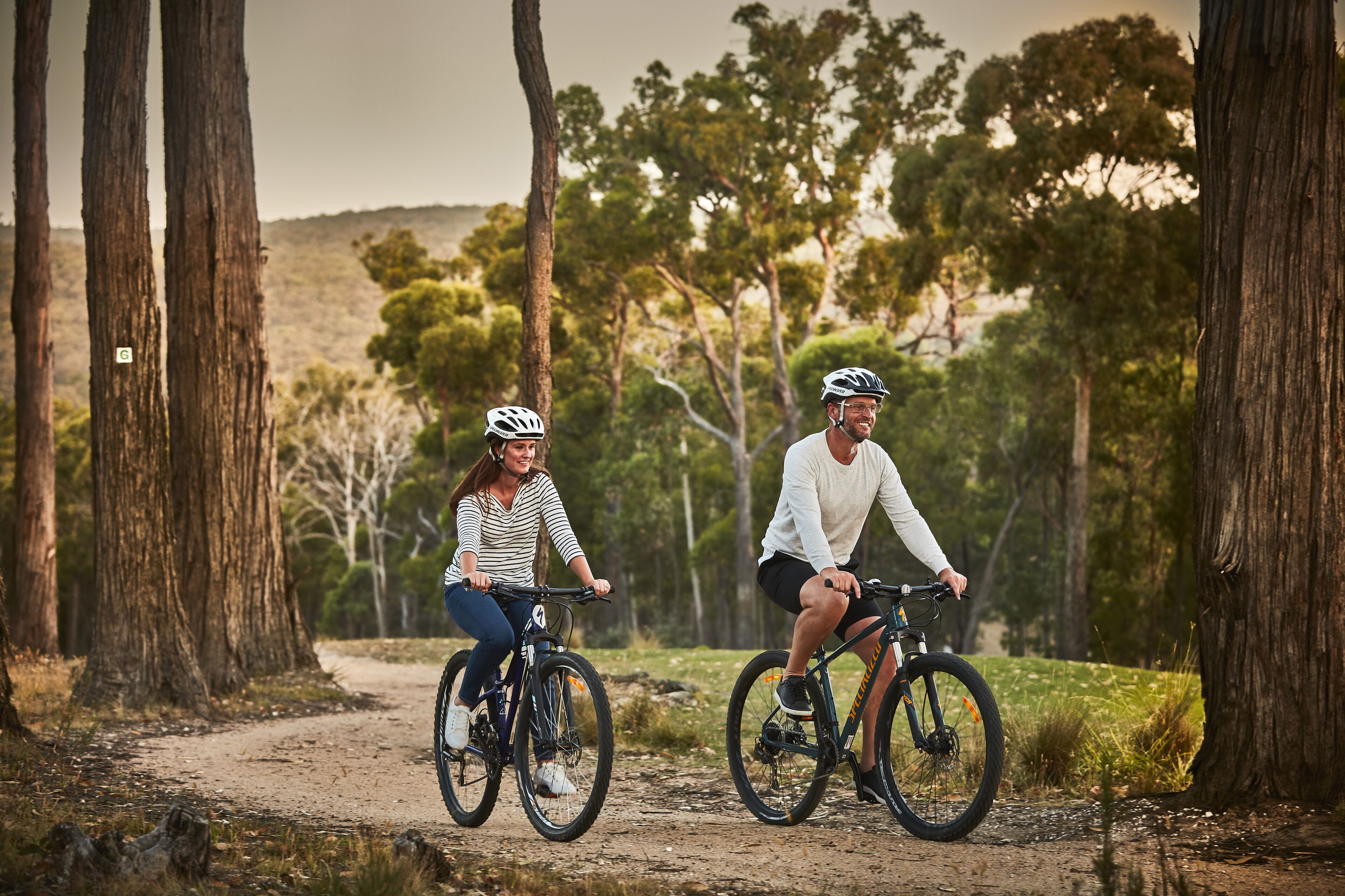 Two people riding bicycles on a dirt path surrounded by trees.