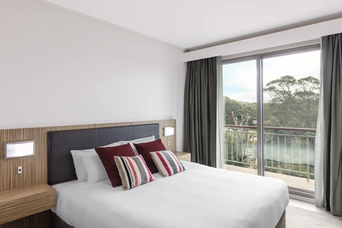 A hotel room with a large bed, a nightstand, a window with curtains, and a view of greenery outside.