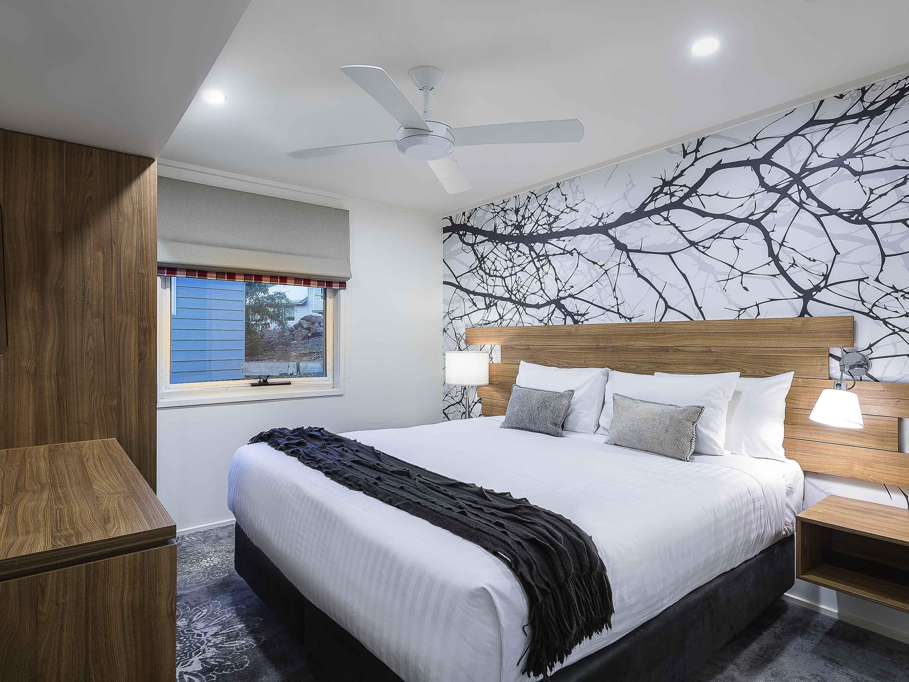 A bedroom with a large bed, a ceiling fan, a window with blinds, and a wall with a tree branch design.