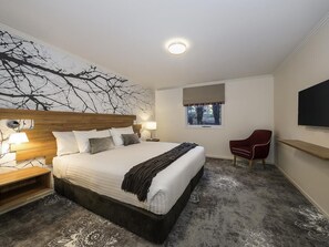 A bedroom with a large bed, bedside tables, a red chair, a window, and a wall-mounted TV.