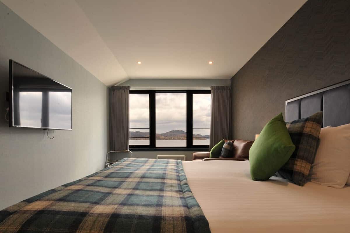 A hotel room with a large bed, a sofa, a TV mounted on the wall, and a view of the mountains through the window.