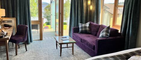 A hotel room with a bed, a sofa, a desk with a chair, and a view of a lake and hills.