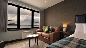 Habitación de hotel con vistas a un lago, un sofá de cuero, una mesita redonda pequeña, una cama con manta escocesa y una mesita de noche con lámpara.