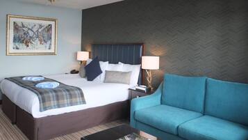 A hotel room with a bed, two bedside lamps, a blue sofa, a coffee table, and a framed artwork on the wall.