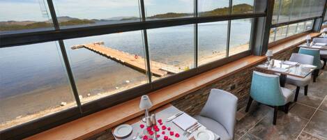 A restaurant with a view of a dock and water, featuring a table set with red rose petals, white plates, and silverware.