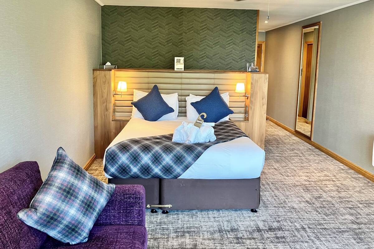A hotel room with a bed, a purple armchair, a ceiling fan, and a herringbone-patterned wall.