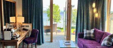 Habitación de hotel con escritorio, sillón morado y sofá con vista al lago.