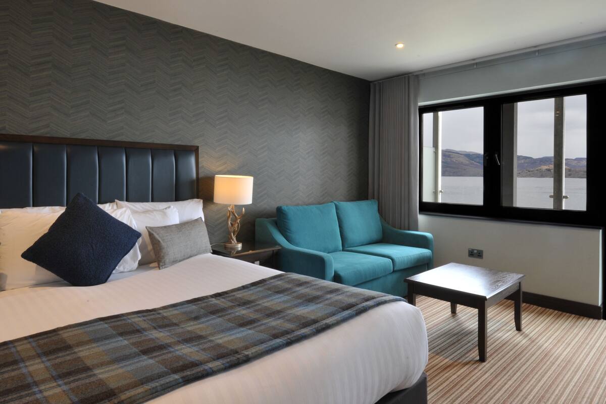 A hotel room with a large bed, a sofa, a small table, and a view of the water through a window.