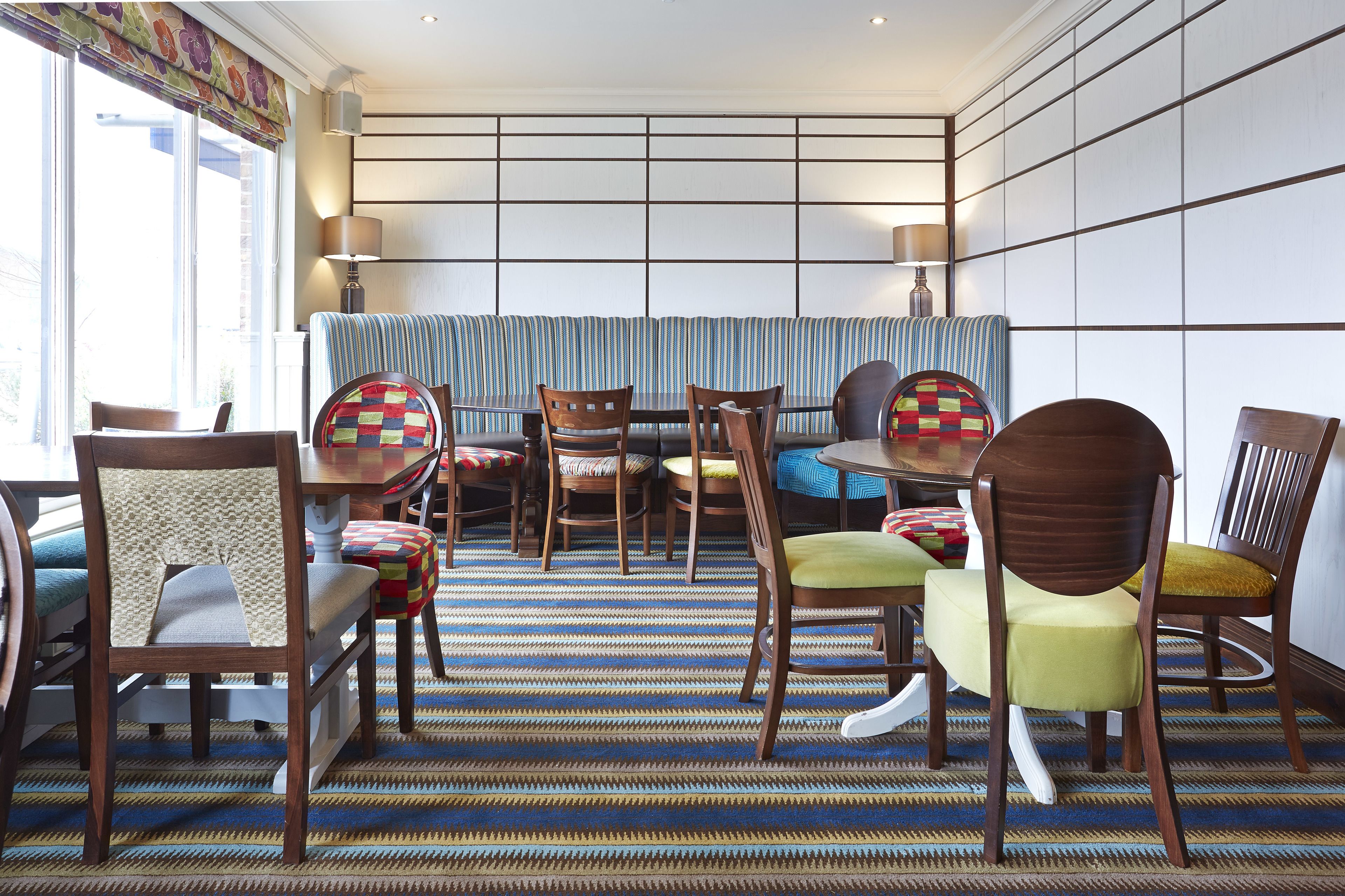 A dining area with a striped bench, wooden chairs with colorful cushions, and a patterned carpet.