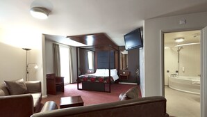 Ein Hotelzimmer mit Himmelbett, einer Sitzecke inklusive Sofa und Sesseln, einem großen Spiegel und einem separaten Badezimmer mit Dusche und Badewanne.