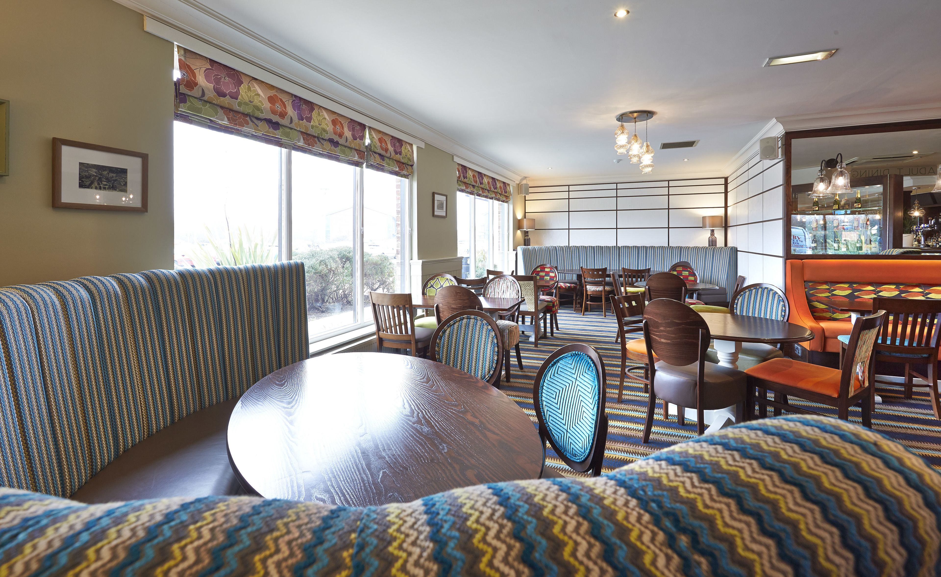 Un comedor con varias mesas y sillas, una zona de bar con mostrador y taburetes, y amplias ventanas con cortinas.