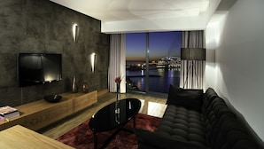 A modern living room with a flat-screen TV, wooden console, black sofa, and a view of a cityscape at dusk. - Ilayda Avantgarde Hotel (Kusadasi)