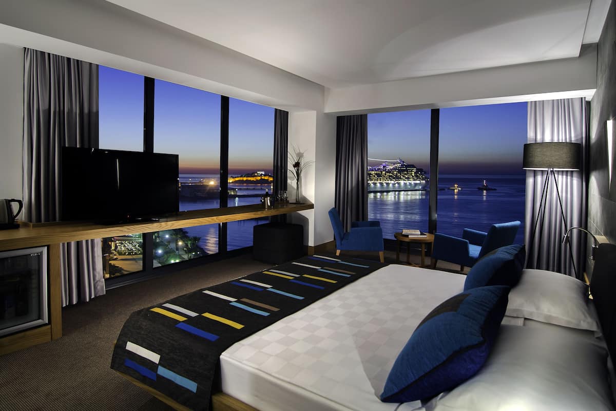 A modern hotel room with a large bed, a TV, a desk with a computer, and a view of the sea.