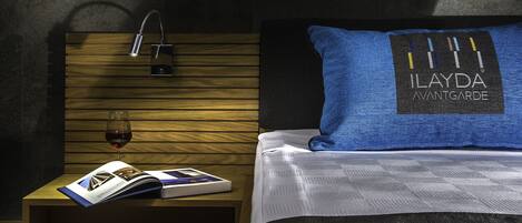 Ein Hotelzimmer mit einem Holzkopfteil, einem blauen Kissen mit dem Markennamen, einem Nachttisch mit einer Zeitschrift und einem Glas Wein sowie einem modernen Wasserhahn.