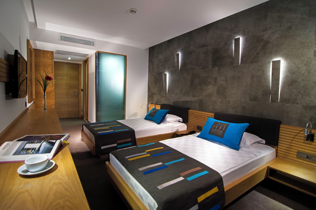A hotel room with two beds, a wooden desk, a TV, and a large window.