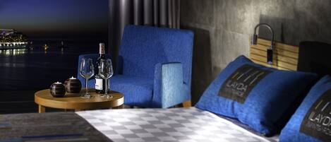 A hotel room with a checkered bedspread, a blue upholstered chair, a small round table with a bottle and glasses, and a view of the sea.