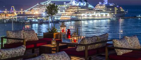 Ein Deck mit roter Polsterung und Laternen, mit Blick auf ein Kreuzfahrtschiff bei Nacht.