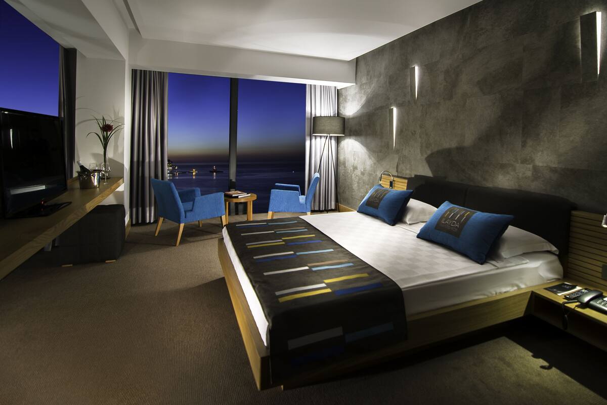 A modern hotel room with a large bed, a desk, a TV, and a view of the ocean.
