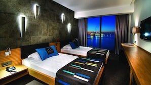 A hotel room with two beds, a wooden desk, a TV, and a view of the city at dusk. - Ilayda Avantgarde Hotel (Kusadasi)