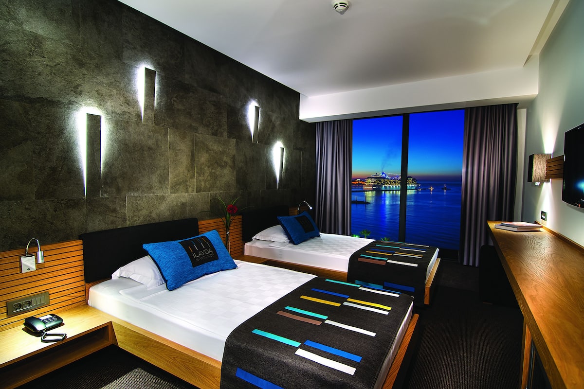 A hotel room with two beds, a wooden desk, a TV, and a view of the city at dusk.