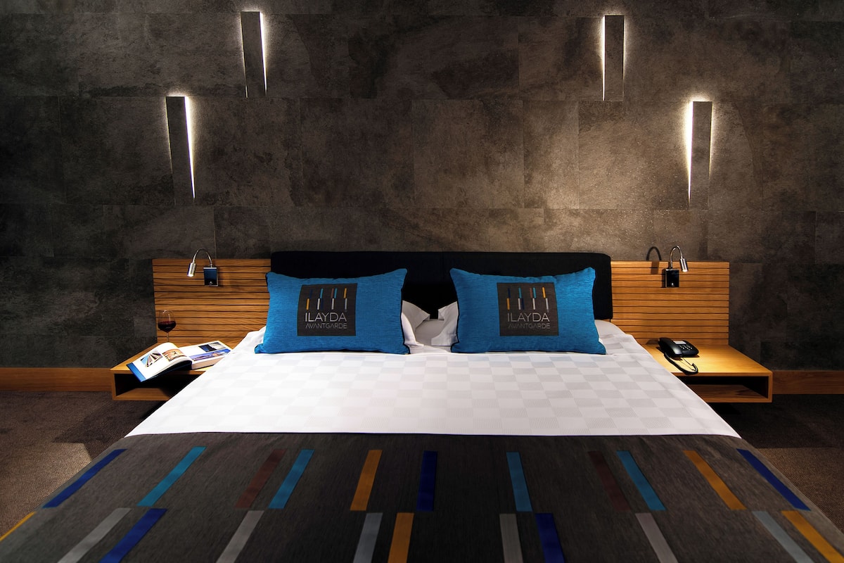 A modern hotel room with a large bed, two blue pillows, a wooden headboard, a bedside table with a phone, and a magazine.