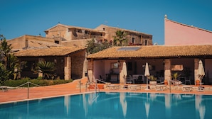 Una piscina con sillas de descanso y sombrillas, rodeada de edificios con techos de tejas rojas y paredes de piedra.