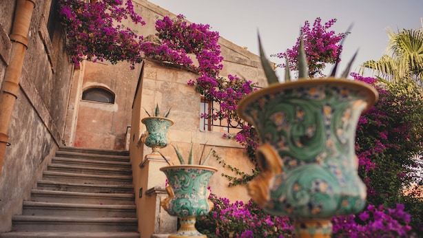 Una escalera conduce a un edificio con fachada de piedra, rodeado de flores moradas vibrantes y jarrones decorativos.