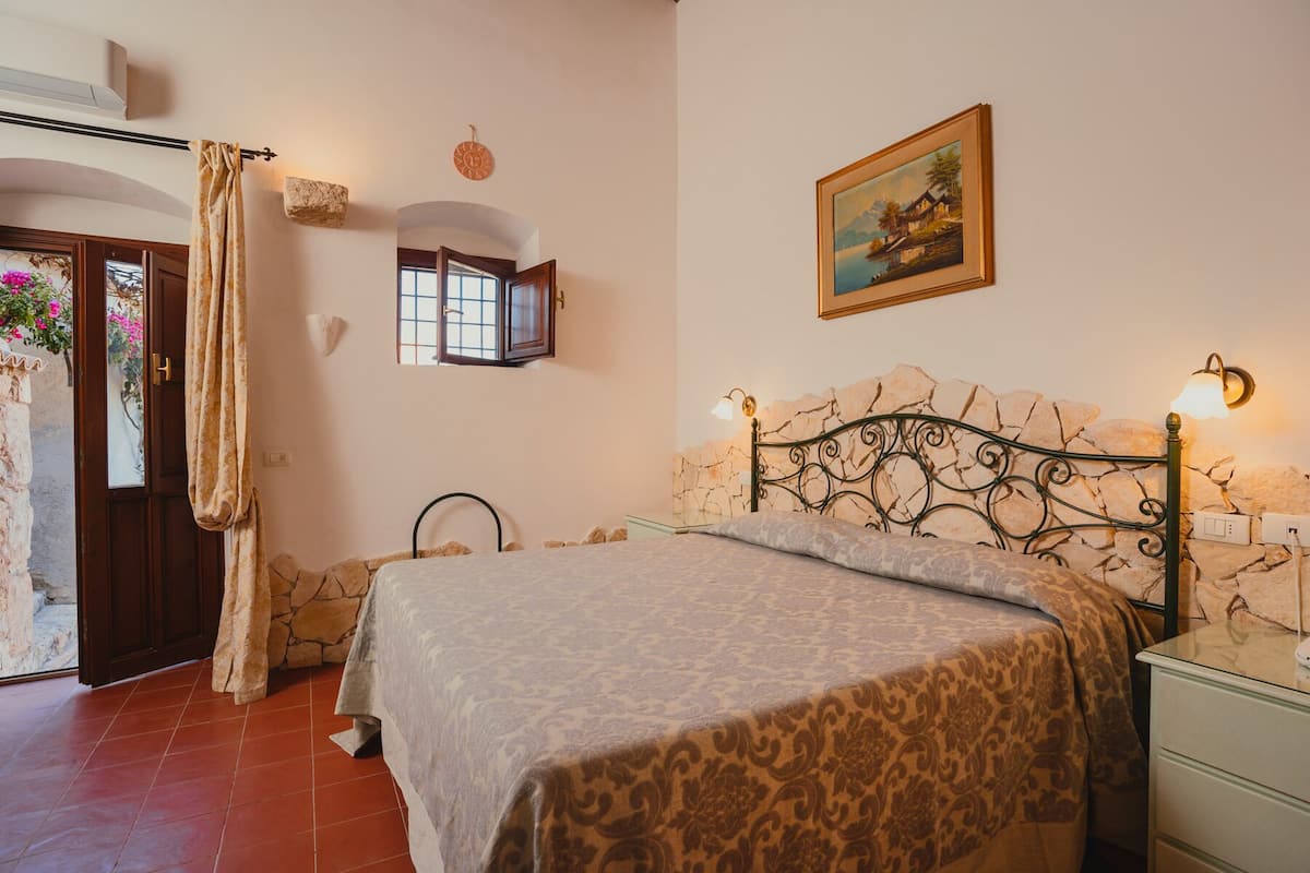 A bedroom with a bed, a painting, a stone wall, a window, and a door.