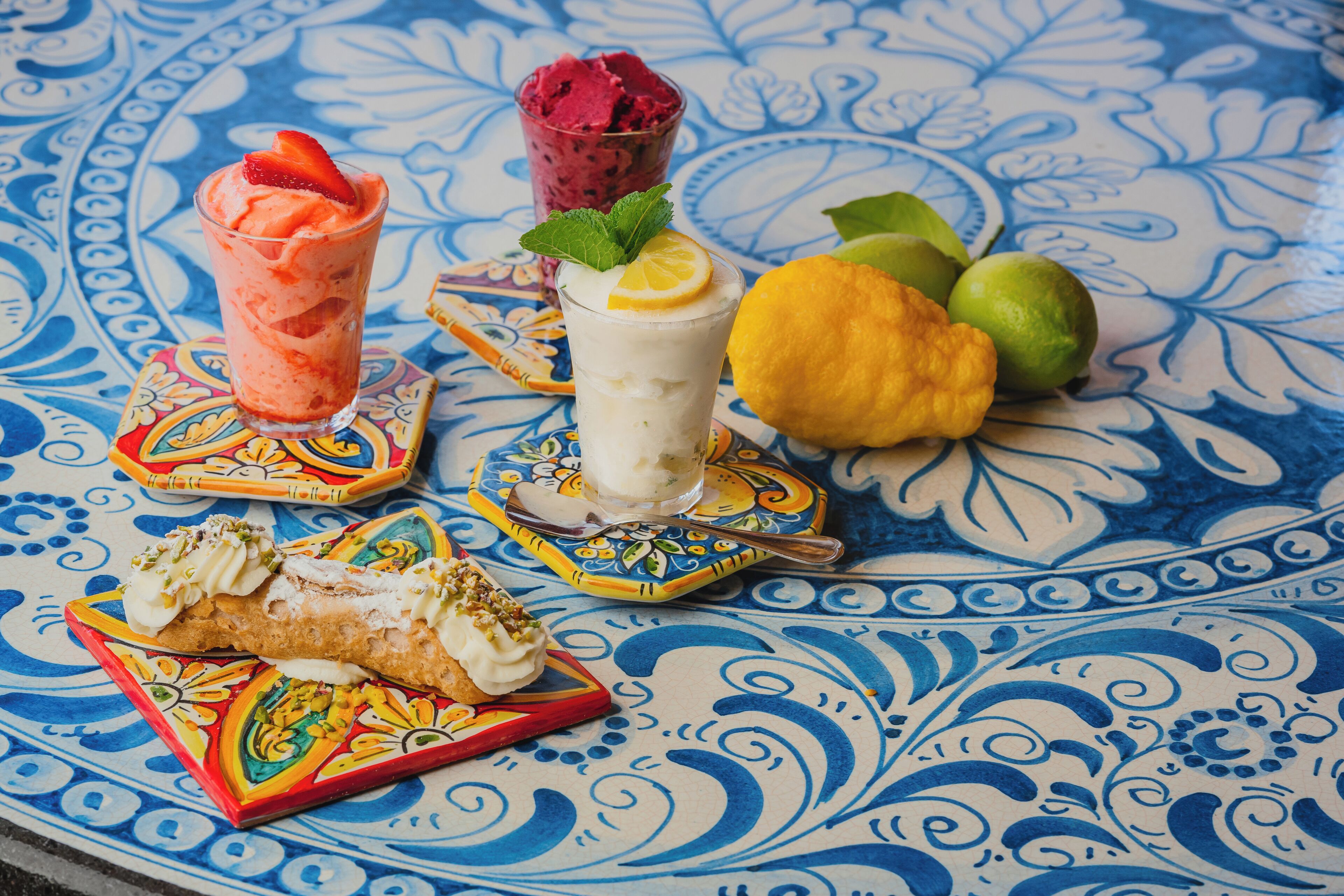 A table with a blue and white patterned tablecloth, a glass of strawberry-flavored drink, a glass of white drink with a lemon slice, a piece of cake, and some fruit.