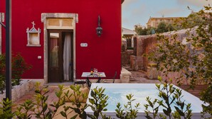 Un edificio rojo con una puerta, una mesa pequeña con sillas y una piscina al frente.