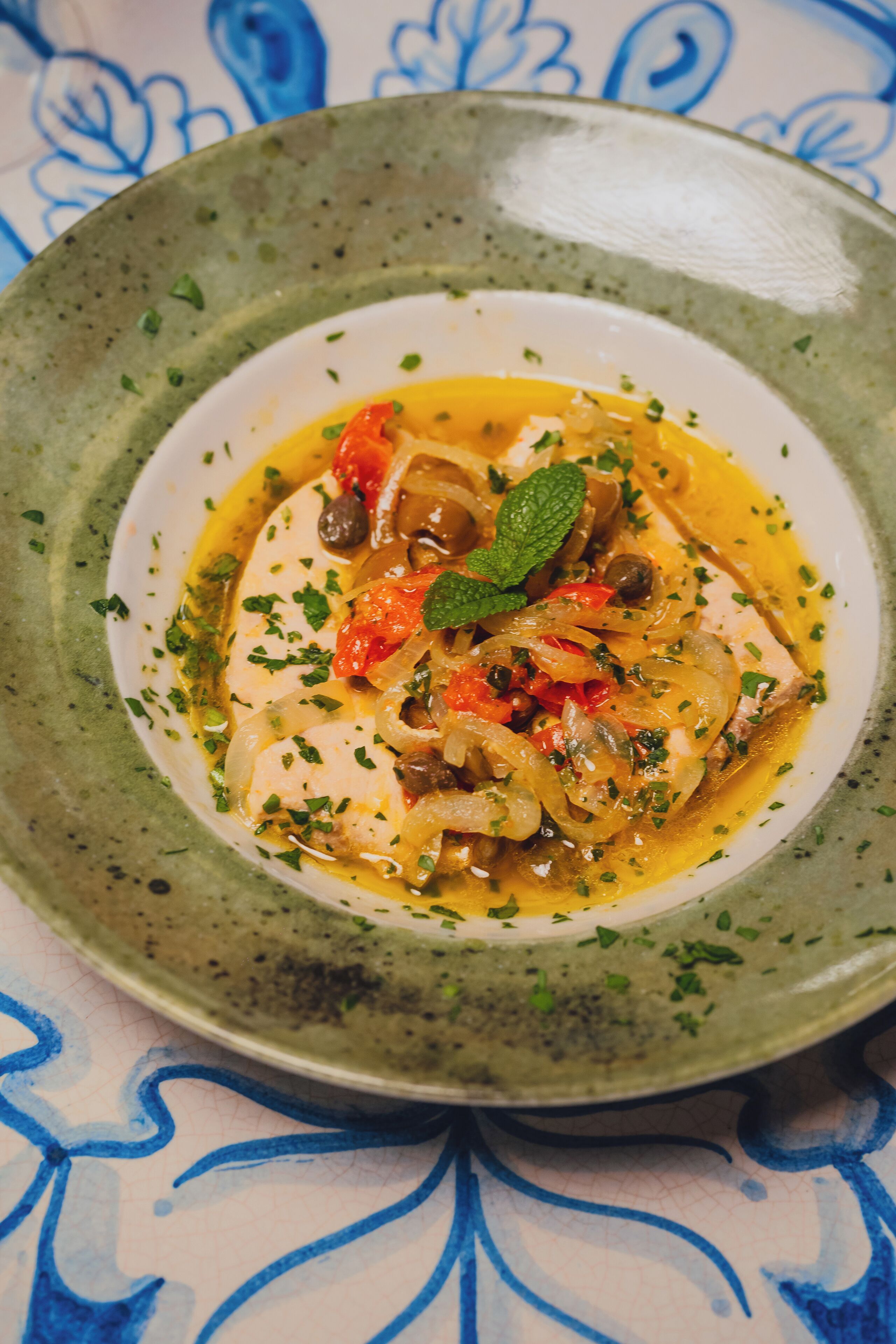 A plate of food with a green rim, garnished with herbs and olives, on a table with a blue and white pattern.