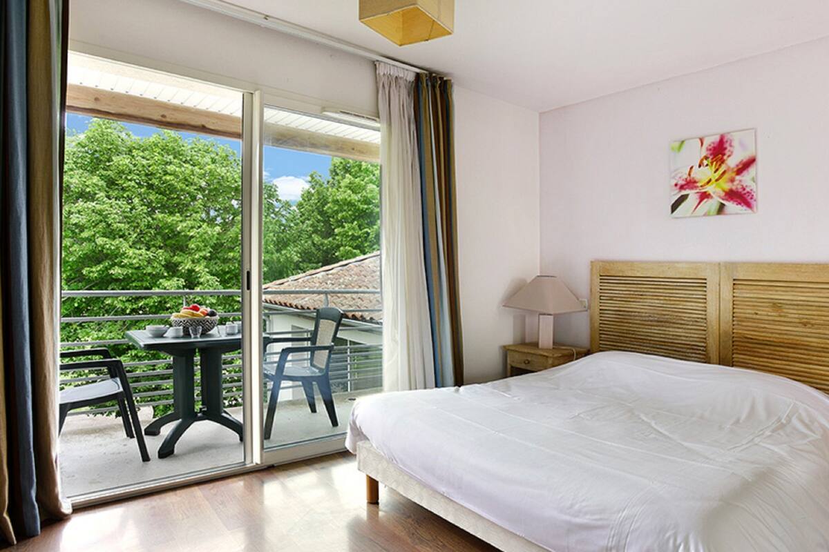 Une chambre d’hôtel avec un lit, une table de chevet, une lampe, un tableau, un balcon avec des chaises et une table avec des fruits.