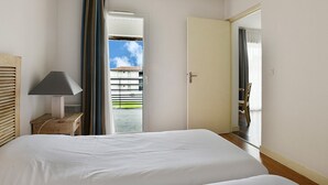 Une chambre d’hôtel avec un lit, une table de chevet avec une lampe, une porte donnant sur un balcon et une vue sur les bâtiments environnants.